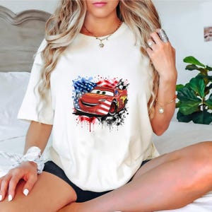 May include: A white short-sleeved t-shirt featuring a graphic print of a red race car with a smiling face. The car is layered over a watercolor-style American flag design with blue stars and red and white stripes, accented by black checkered patterns.
