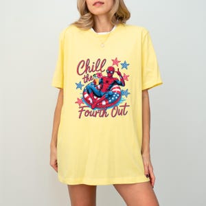 May include: A pale yellow short-sleeve t-shirt featuring a graphic of a superhero in a red and blue suit relaxing in a patriotic red, white, and blue inner tube while holding a drink. The text reads Chill the Fourth Out with star accents.