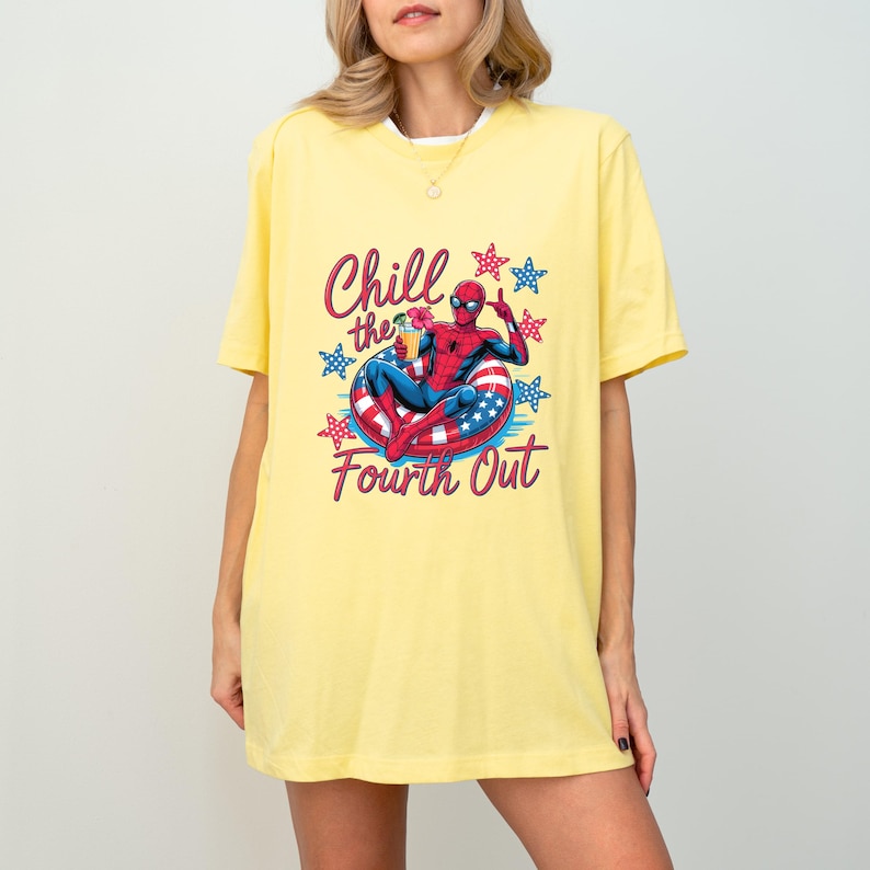 May include: A pale yellow short-sleeve t-shirt featuring a graphic of a superhero in a red and blue suit relaxing in a patriotic red, white, and blue inner tube while holding a drink. The text reads Chill the Fourth Out with star accents.