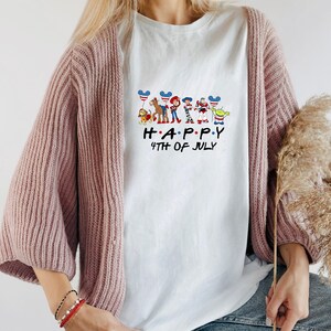 May include: A white t-shirt featuring a graphic of various animated characters holding balloons with American flag patterns. Below the characters, the text reads "H A P P Y 4TH OF JULY" in black lettering. The shirt is worn under a dusty pink knit cardigan.