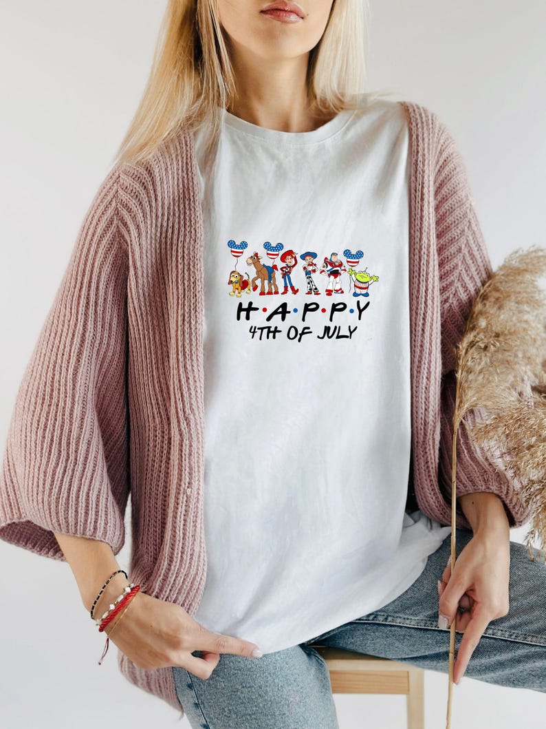 May include: A white t-shirt featuring a graphic of various animated characters holding balloons with American flag patterns. Below the characters, the text reads "H A P P Y 4TH OF JULY" in black lettering. The shirt is worn under a dusty pink knit cardigan.