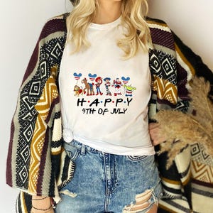 May include: A white short-sleeved t-shirt featuring a graphic of cartoon characters holding balloons with American flag patterns, with the text "H.A.P.P.Y 4TH OF JULY" printed in black below the characters, worn with blue denim shorts and a patterned cardigan.