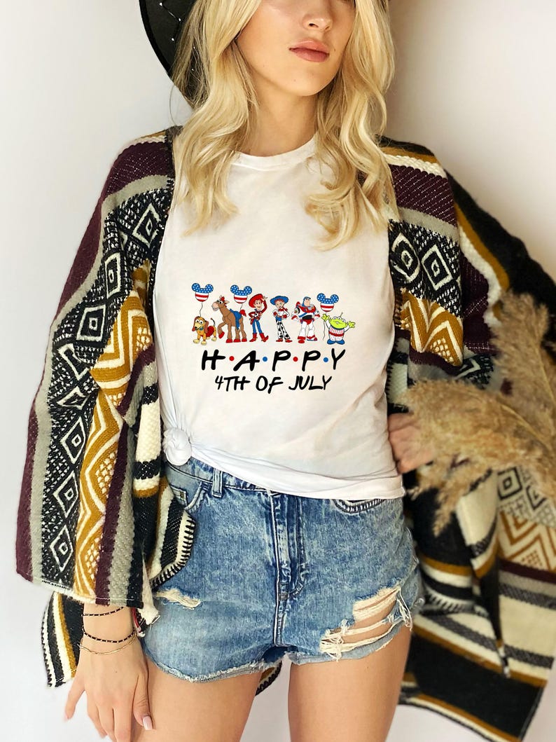 May include: A white short-sleeved t-shirt featuring a graphic of cartoon characters holding balloons with American flag patterns, with the text "H.A.P.P.Y 4TH OF JULY" printed in black below the characters, worn with blue denim shorts and a patterned cardigan.