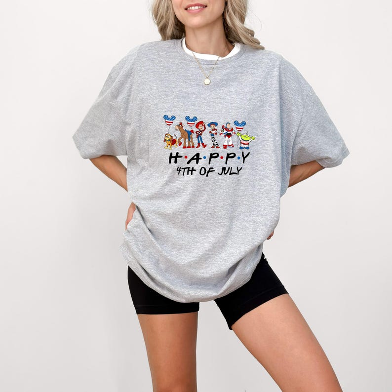 May include: A heather gray short-sleeve t-shirt featuring a graphic of Toy Story characters holding American flag-patterned mouse ears. Below the characters, the text reads HAPPY 4TH OF JULY in black lettering. The shirt has a crew neckline.