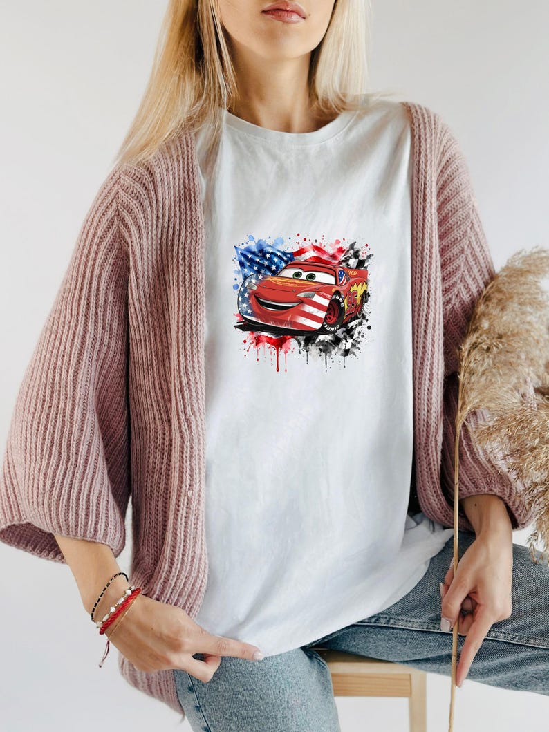 May include: A white t-shirt featuring a graphic print of a red race car with a smiling face, overlaid with a watercolor-style American flag design, black paint splatters, and a checkered flag pattern on the right side of the car illustration.