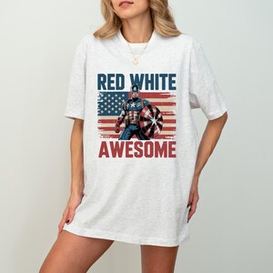 May include: A light heather gray short-sleeve t-shirt featuring a graphic of Captain America holding a shield in front of a distressed American flag. The text RED WHITE is printed in blue above the graphic, and AWESOME is printed in red below.