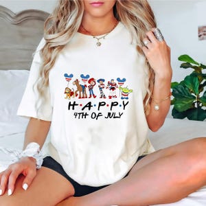 May include: A cream-colored short-sleeve t-shirt featuring a graphic of various animated characters, including a dog, a horse, and space-themed figures, holding balloons with American flag patterns. The text reads HAPPY 4TH OF JULY.