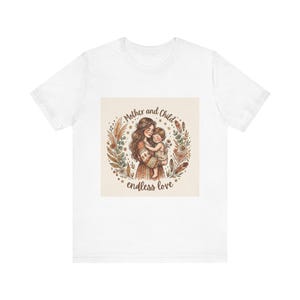 Könnte beinhalten: Weißes T-Shirt mit einer Aquarellillustration einer Mutter und eines Kindes, die in einem floralen Kranz umarmt sind. Der Text "Mother and Child endless love" befindet sich über und unter dem Bild. Das Design ist in warmen Erdtönen gehalten.