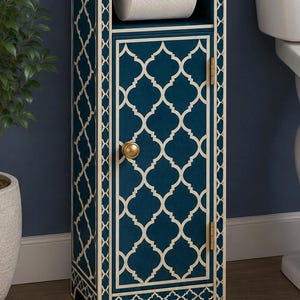 May include: A tall, narrow, dark blue bathroom storage cabinet featuring a white quatrefoil inlay pattern. The unit includes an open top shelf holding a roll of toilet paper on a gold rod and a lower cabinet door with a round gold knob.