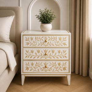 May include: A white three-drawer nightstand featuring gold-toned trim and carved floral patterns on each drawer front. Each drawer is equipped with a single round gold knob. A small potted green plant sits on the flat top surface of the unit.