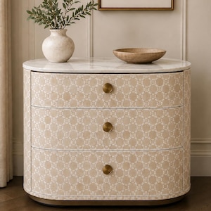 May include: A curved three-drawer chest featuring a light beige and white geometric pattern on the drawer fronts, a white marble top, and three circular brass-toned knobs. A speckled ceramic vase with green foliage and a shallow bowl sit on top.