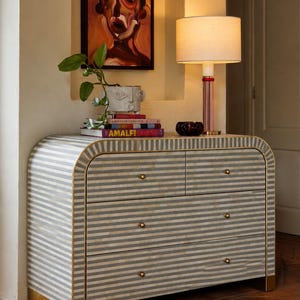 May include: A curved chest of drawers featuring a gray and white striped pattern with gold-toned trim and hardware. The piece has two small top drawers and two full-width bottom drawers, all accented with small, round, metallic knobs.
