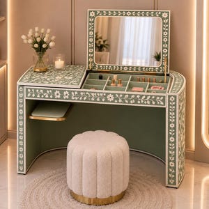 Puede incluir: Un tocador verde salvia con incrustaciones florales blancas, que cuenta con un espejo abatible, compartimentos de almacenamiento internos y un taburete cilíndrico capitoné de color crema a juego con base dorada, sobre una alfombra redonda.