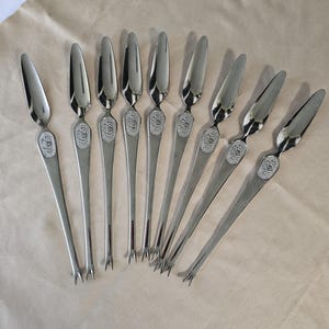 May include: A set of nine silver-toned metal seafood forks, each featuring a polished, pointed bowl at the top and a two-pronged tip at the bottom. The handle of each fork is adorned with a small, circular embossed emblem depicting a crab.