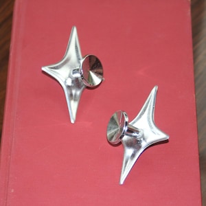 May include: Two silver-colored metal drawer knobs with a four-pointed star shape. Each knob features a central circular post and a flat, reflective surface that tapers into sharp, elongated points at the top, bottom, left, and right sides.