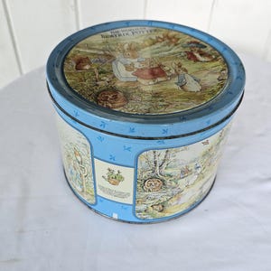 May include: A round, light blue metal storage tin featuring illustrations from The World of Beatrix Potter. The lid displays Peter Rabbit and other characters in a garden setting, while the sides show various scenes and text in panels.