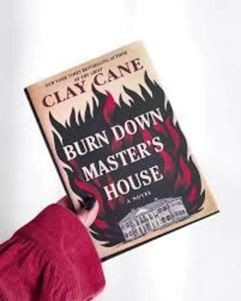 Puede incluir: Portada del libro con el t&iacute;tulo "Burn Down Master's House" de Clay Cane. La portada presenta un fondo negro con llamas rojas y una ilustraci&oacute;n blanca de una casa. El libro est&aacute; sostenido por una persona que lleva una camisa roja.