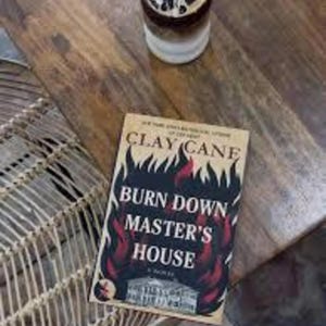 Puede incluir: Un libro titulado "BURN DOWN MASTER'S HOUSE" de Clay Cane, con un dise&ntilde;o ardiente, descansa sobre una mesa de madera. Un vaso de caf&eacute; helado est&aacute; cerca. La escena est&aacute; ambientada en una mesa de madera con una silla de mimbre visible.