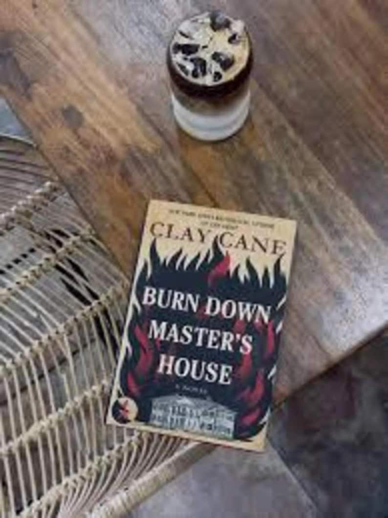 Puede incluir: Un libro titulado "BURN DOWN MASTER'S HOUSE" de Clay Cane, con un dise&ntilde;o ardiente, descansa sobre una mesa de madera. Un vaso de caf&eacute; helado est&aacute; cerca. La escena est&aacute; ambientada en una mesa de madera con una silla de mimbre visible.