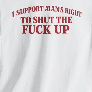 Può includere: Una t-shirt bianca a maniche corte con testo rosso stampato sul petto che recita: I SUPPORT MAN'S RIGHT TO SHUT THE FUCK UP. Il capo presenta uno scollo rotondo e una vestibilità standard, con le lettere ben visibili sul tessuto bianco.