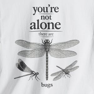 Peut inclure: Un t-shirt blanc à manches courtes avec une illustration graphique noire représentant trois libellules. Le texte imprimé sur le devant indique, you're not alone there are bugs, disposé de manière centrée au-dessus et en dessous des insectes.