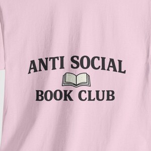 Può includere: Una t-shirt a maniche corte color rosa chiaro con la scritta nera ANTI SOCIAL BOOK CLUB stampata sul petto. Tra le parole ANTI SOCIAL e BOOK CLUB si trova un piccolo e semplice disegno a linee nere di un libro aperto con pagine visibili.