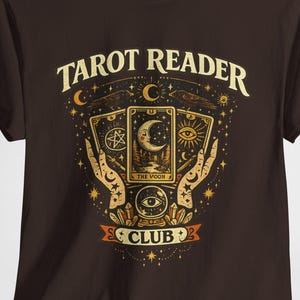 Può includere: Una t-shirt a maniche corte marrone scuro con una stampa grafica recante il testo "TAROT READER CLUB". Il design include tre carte dei tarocchi, due mani, una sfera di cristallo, stelle e lune crescenti nei toni del giallo, arancione e bianco.