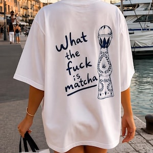 Könnte beinhalten: Ein weißes kurzärmeliges T-Shirt mit einer dunkelblauen Grafik auf der Rückseite. Die Grafik zeigt eine stilisierte Abbildung einer Tequila-Flasche und den Text "What the fuck is matcha" in einer lässigen, handschriftlichen Schriftart.