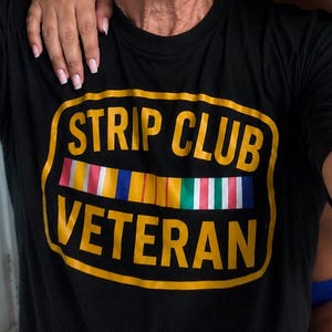 Op de afbeelding: Een zwart T-shirt met korte mouwen en een geel omrande afbeelding met de tekst STRIP CLUB VETERAN in dikke gele letters. Tussen de twee tekstregels staat een horizontale rij kleurrijke verticale strepen die doen denken aan militaire onderscheidingen.