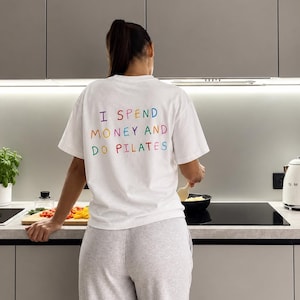 Può includere: Una t-shirt bianca a maniche corte con la scritta "I SPEND MONEY AND DO PILATES" stampata sul retro in lettere multicolori. La maglietta è abbinata a pantaloni della tuta grigio chiaro, creando un outfit casual per la vita quotidiana.