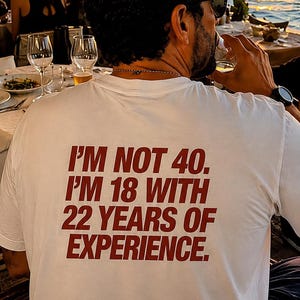 Könnte beinhalten: Ein weißes kurzärmeliges T-Shirt mit dunkelrotem Text auf der Rückseite, der lautet: I'M NOT 40. I'M 18 WITH 22 YEARS OF EXPERIENCE. Das Shirt wird von einer Person getragen, die von hinten zu sehen ist, wobei der Fokus auf dem Aufdruck liegt.