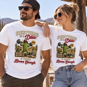 Può includere: Una t-shirt bianca con la grafica di un cactus verde che solleva un bilanciere in un deserto. Il testo recita Everything is a Dildo, No Pain No Gain e If You're Brave Enough. Il design include un sole e dei pesi da palestra.
