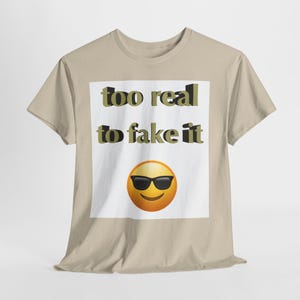 Può includere: Una maglietta beige con la scritta "too real to fake it" in lettere nere e verde oliva. Sotto il testo c'è un'emoji gialla con occhiali da sole neri. La maglietta è realizzata in materiale morbido.