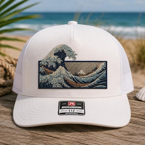May include: A white trucker hat featuring a rectangular patch with an embroidered depiction of a large ocean wave and a mountain. The hat has a structured front panel, a curved brim, and a black sticker with the text Richardson 112 Adjustable.