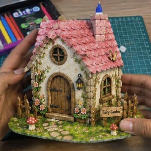 Peut inclure: Une maison miniature avec un toit en bardeaux roses, des murs en pierre et une porte en bois. La structure comprend une cheminée, une petite lanterne et des vignes grimpantes avec des fleurs roses. Une clôture en bois entoure la maison, avec deux champignons rouges et un panneau indiquant Fairy Garden sur la base verte.