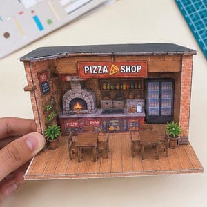 May include: A miniature paper craft model of a pizza shop featuring a brick exterior, a stone oven with a fire graphic, two wooden tables with chairs, a menu board, a refrigerator, and a sign that reads PIZZA SHOP with a slice icon.