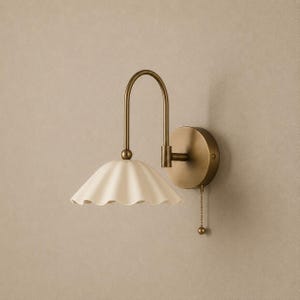Peut inclure: Une applique murale dotée d'un bras incurvé en laiton antique et d'une plaque murale circulaire. Le luminaire comprend un abat-jour festonné blanc cassé et un petit interrupteur à tirette suspendu à la base, offrant une solution d'éclairage.