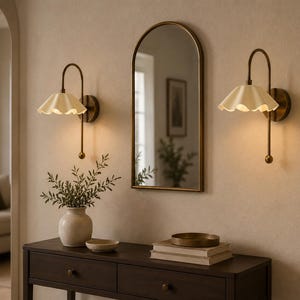 May include: Two wall-mounted lamps with brass-toned curved arms and cream-colored scalloped shades, positioned on either side of an arched mirror with a thin brass frame, all mounted above a dark wooden console table with two drawers.