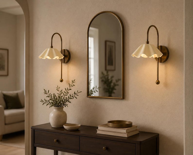 May include: Two wall-mounted lamps with brass-toned curved arms and cream-colored scalloped shades, positioned on either side of an arched mirror with a thin brass frame, all mounted above a dark wooden console table with two drawers.