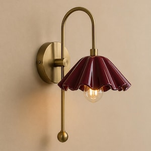Peut inclure: Une applique murale composée d'un bras en métal doré avec une base circulaire et d'un abat-jour en céramique cannelée de couleur bordeaux profond. Le luminaire comprend une ampoule apparente diffusant une lumière chaude sous l'abat-jour.