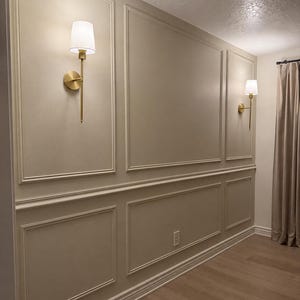 K&ouml;nnte beinhalten: Eine Wand mit beigefarbener Wandvert&auml;felung mit rechteckigen Zierleisten und zwei Wandleuchten aus Messing mit wei&szlig;en Stoffschirmen. Die Wand ist in einem einheitlichen Beigeton gestrichen, erg&auml;nzt durch hellen Holzboden und beige Vorh&auml;nge.