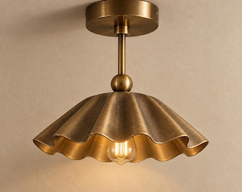 May include: A semi-flush mount ceiling light fitting with a brushed brass finish. The lamp features a wide, scalloped metal shade with a wavy edge and a central stem connected to a circular canopy, housing a single exposed Edison-style light bulb.
