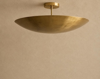 Może przedstawiać: Lampa sufitowa z szerokim, płytkim kloszem w kształcie misy wykonanym ze szczotkowanego mosiądzu. Oprawa jest zawieszona na krótkim, cienkim pionowym pręcie połączonym z okrągłą podsufitką, która jest przymocowana do powierzchni sufitu.