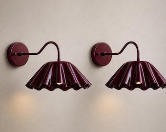 Może przedstawiać: Para pasujących lamp ściennych w kolorze głębokiego burgundu. Każda lampa składa się z okrągłej podstawy montażowej, wygiętego ramienia oraz szerokiego, falistego i żłobkowanego klosza, który kieruje światło w dół z centralnego gniazda.