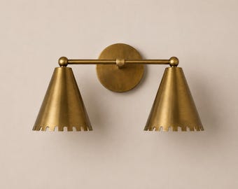 May include: A wall-mounted light fitting with a brushed brass finish, featuring a circular backplate and a horizontal bar supporting two downward-facing conical shades. Each shade has a decorative notched edge along the bottom rim.
