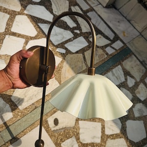 May include: A wall-mounted light fixture featuring a curved antique brass arm and a circular mounting plate. The lamp includes a pale cream-colored, fluted metal shade with a scalloped edge and a thin, straight brass rod extending downward.