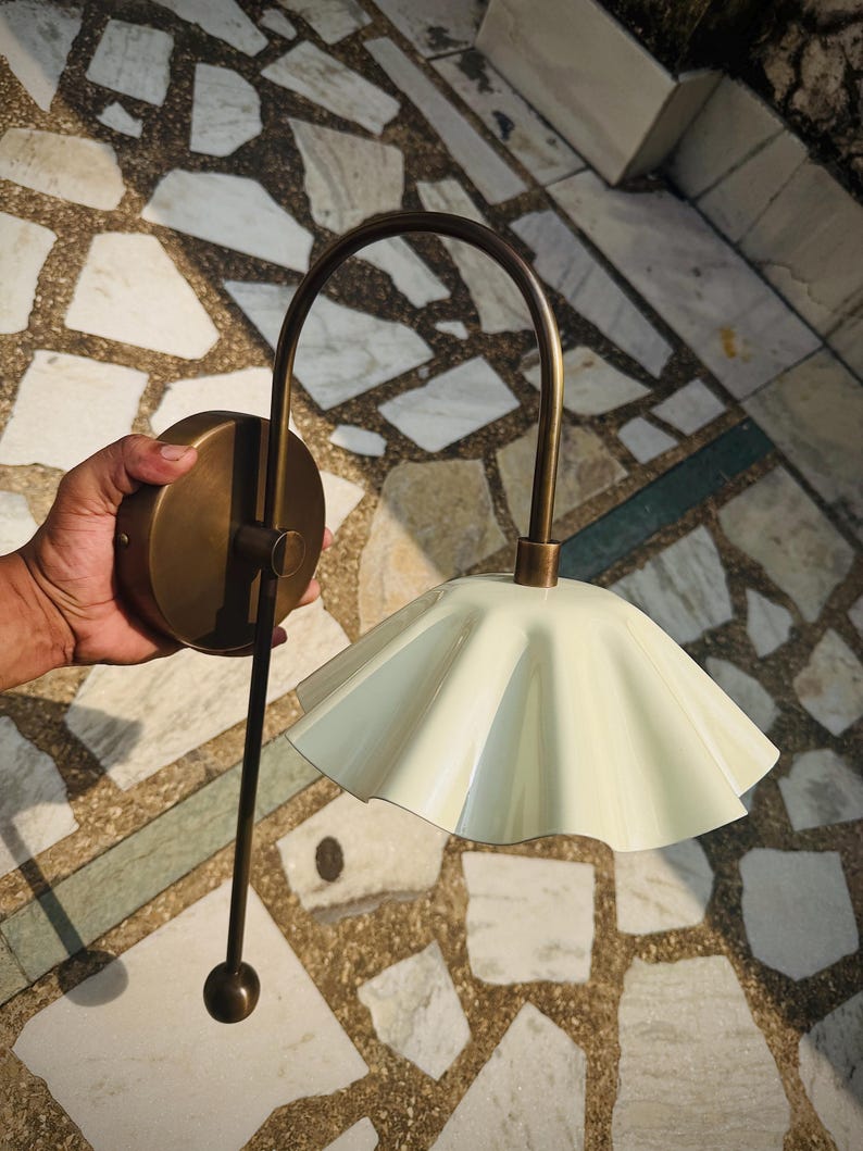 May include: A wall-mounted light fixture featuring a curved antique brass arm and a circular mounting plate. The lamp includes a pale cream-colored, fluted metal shade with a scalloped edge and a thin, straight brass rod extending downward.