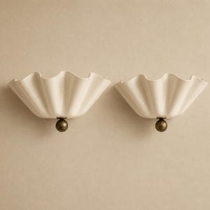 May include: A pair of wall lights featuring off-white, ruffled, fan-shaped shades with a smooth, glossy finish. Each shade is attached to a central, spherical, dark metallic bronze-toned base that connects to the wall mounting point.