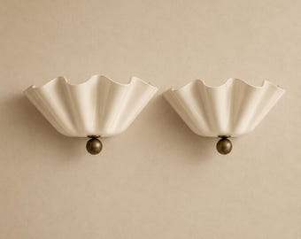 May include: A pair of wall lights featuring off-white, ruffled, fan-shaped shades with a smooth, glossy finish. Each shade is attached to a central, spherical, dark metallic bronze-toned base that connects to the wall mounting point.