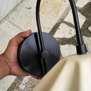 May include: A wall-mounted light fixture featuring a circular matte black base and a curved black metal arm that supports a wide, fluted, cream-colored lampshade. The design includes a central black connector piece where the arm meets the base.
